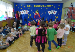 Dekoracja Andrzejkowa, przy niej nauczycielka przebrana za Cygankę. 7 dzieci stoi na srodku sali, pozstałe dzieci siedzą po bokach.